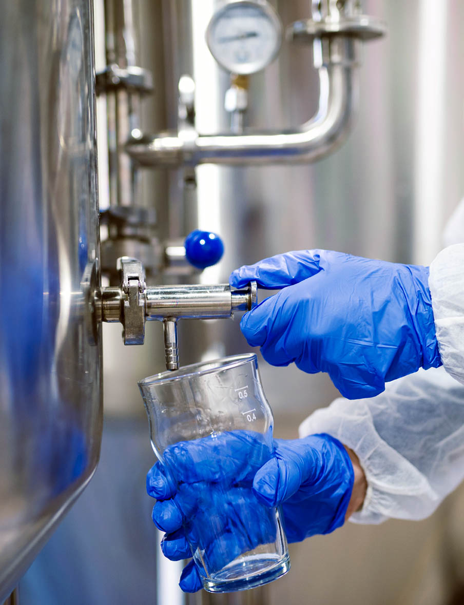 Close up view of hands in protective gloves opening tap valve and filling glass with samples from industrial machine reservoir.